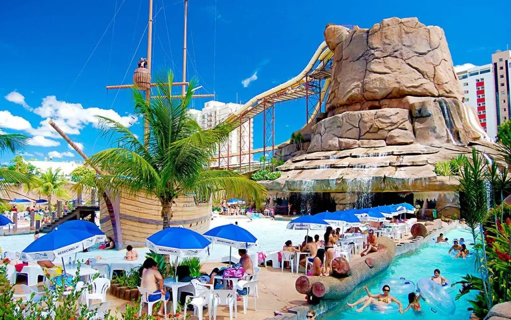 Parque aquático com piscina, escorregadores, grande rocha artificial, pessoas se divertindo e guarda-sóis azuis, em dia ensolarado.