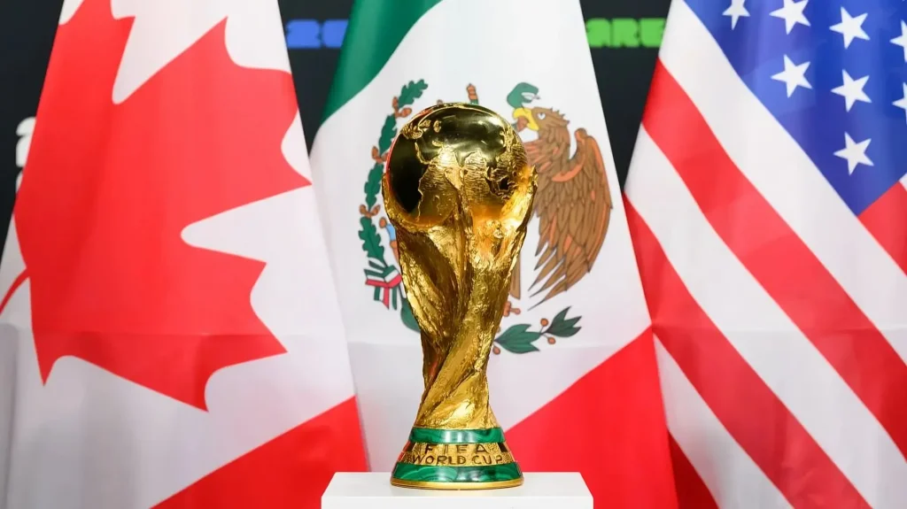 Troféu dourado da Copa do Mundo FIFA posicionado em destaque sobre um suporte branco. Ao fundo, ocupando toda a largura da imagem, estão as bandeiras nacionais do Canadá (esquerda), México (centro) e Estados Unidos (direita), representando os países-sede do torneio de 2026.