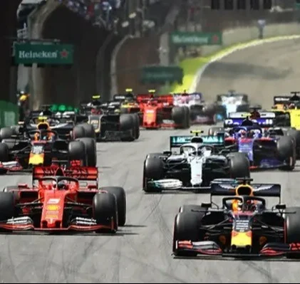 Visão de um pelotão completo de carros de Fórmula 1 correndo juntos em uma curva de um circuito. Em primeiro plano, um carro vermelho da Ferrari, um prata da Mercedes e um azul escuro da Red Bull disputam posição, com o restante do grid seguindo-os de perto.