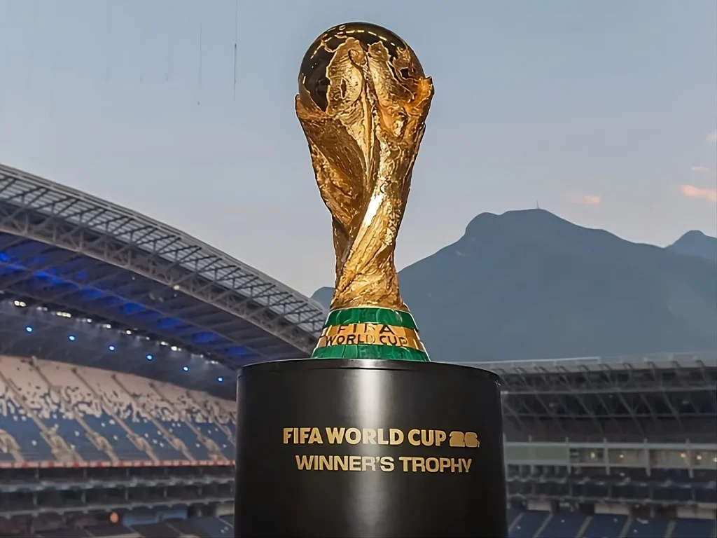 Para acessibilidade e leitura de imagem pelo Google: "Foto em plano próximo do troféu dourado da Copa do Mundo FIFA sobre um pedestal preto com a inscrição 'FIFA World Cup 26 Winner's Trophy'. Ao fundo, vê-se a arquibancada de um estádio com assentos azuis e, no horizonte, grandes montanhas sob um céu de entardecer."