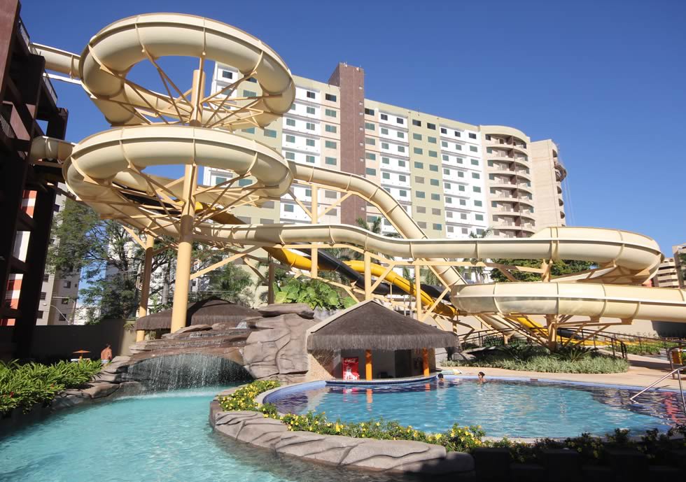 Um complexo de toboáguas grandes e de cor clara, com uma piscina, um bar aquático e um prédio alto e moderno ao fundo.