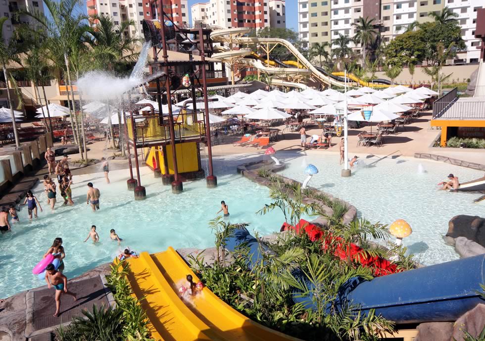 Vista de cima de um parque aquático com várias piscinas e um grande playground aquático amarelo e azul, com crianças e adultos se divertindo. Prédios altos podem ser vistos ao fundo.