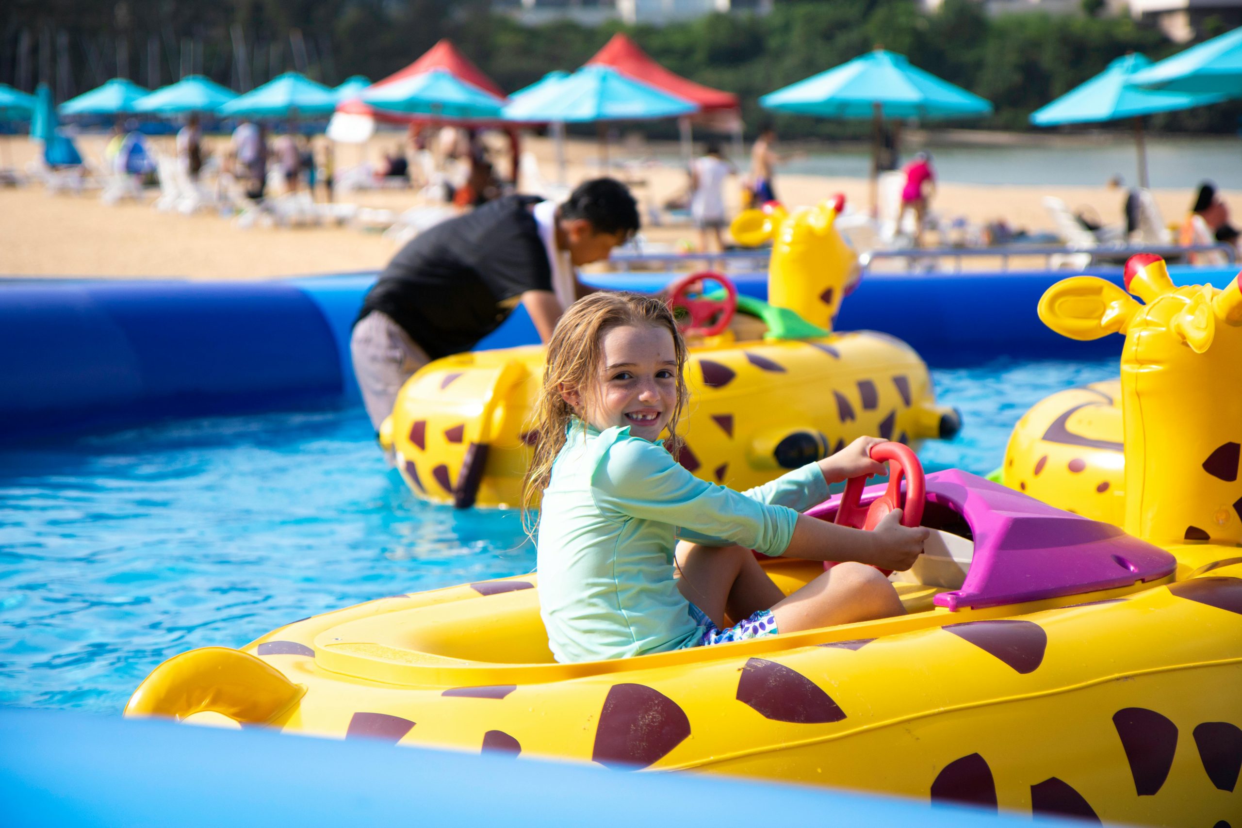 Texto Alternativo (Alt Text):
"Uma menina sorridente brinca em um barco inflável amarelo com formato de girafa em uma piscina infantil azul. Ao fundo, outras pessoas e brinquedos infláveis são visíveis, com cadeiras de praia e guarda-sóis coloridos em uma área de areia."

Descrição:
"A imagem captura um momento de alegria em uma piscina infantil. Uma menina de cabelos claros e camiseta verde de manga comprida está sentada em um barco inflável amarelo, decorado com manchas marrons como uma girafa e detalhes em roxo. Ela segura um volante vermelho e sorri para a câmera. A piscina é de cor azul clara e translúcida, com ondulações na superfície. Ao fundo, parcialmente desfocados, podem-se ver outras pessoas interagindo com brinquedos infláveis amarelos em formato de girafa. Além da piscina, há uma área de areia com diversas cadeiras de praia e guarda-sóis coloridos em tons de azul, vermelho e branco, sugerindo um parque aquático ou área de lazer à beira-mar."

Legenda:
"Dia de diversão na piscina com meu barco-girafa! ☀️ #PiscinaInfantil #BrincadeirasAquáticas #Alegria #Verão"