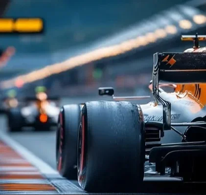 Close-up em ângulo baixo do pneu traseiro de um carro de Fórmula 1 laranja e preto, parado em um pit lane. O foco está nítido no pneu, enquanto um segundo carro e as luzes da garagem estão desfocados ao fundo, criando uma sensação de profundidade.