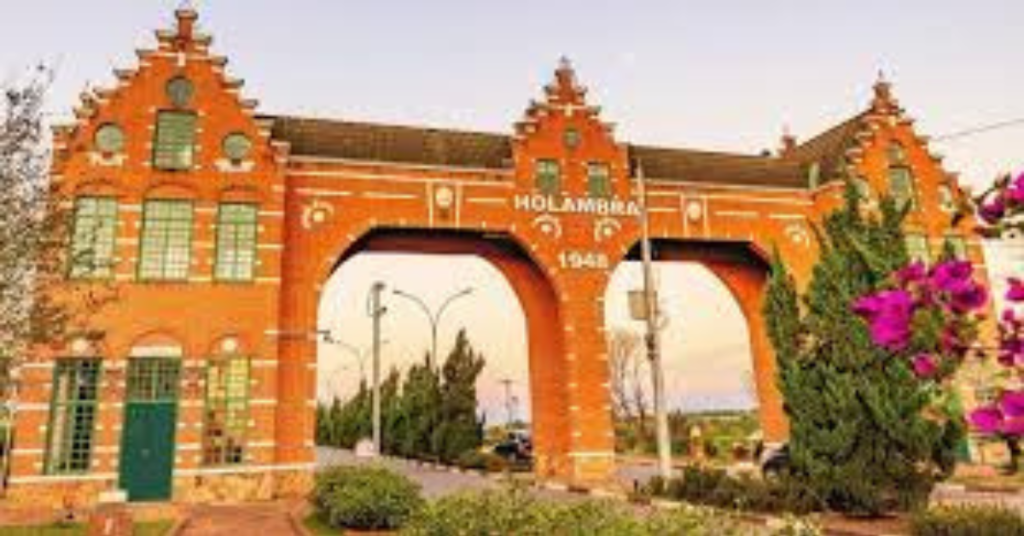 O portal de entrada da cidade de Holambra, uma grande estrutura de tijolos avermelhados com arquitetura holandesa, telhados escalonados e janelas verdes. A palavra "HOLAMBRA" e o ano "1948" estão escritos no topo. Flores rosa e roxas são visíveis no canto direito.