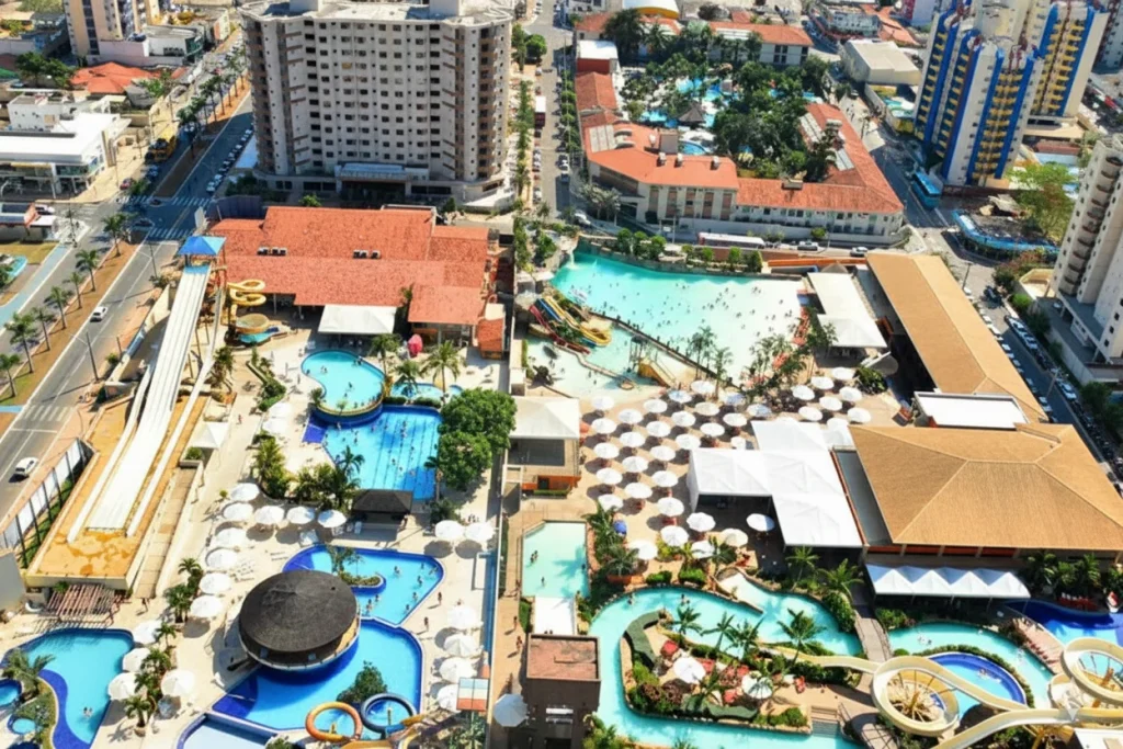 Vista aérea de um extenso complexo de parques aquáticos no centro de Caldas Novas. A imagem mostra um toboágua com múltiplas pistas à esquerda, diversas piscinas lotadas, uma grande área com dezenas de guarda-sóis brancos, e vários hotéis e prédios altos cercando o local por todos os lados.