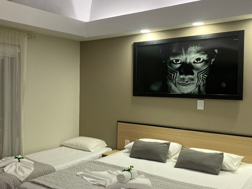 Quarto de hotel no AJ Termas Hotel Iraí com uma cama de casal e uma cama de solteiro auxiliar lado a lado. Na parede acima das camas, há um grande quadro com um retrato fotográfico em preto e branco de um rosto indígena com pintura facial.