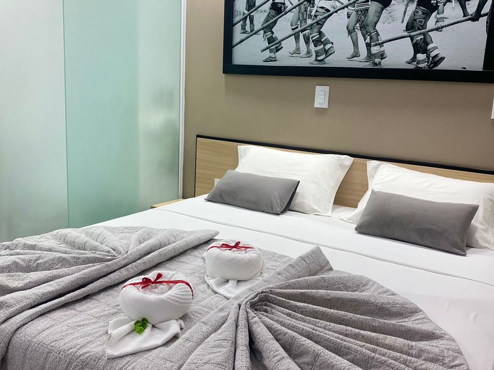 Cama de casal arrumada em um quarto do AJ Termas Hotel Iraí, com lençóis brancos, colcha cinza e duas esculturas de cisnes de toalha decoradas com fitas vermelhas. Ao fundo, uma cabeceira de madeira clara e um quadro artístico em preto e branco na parede.
