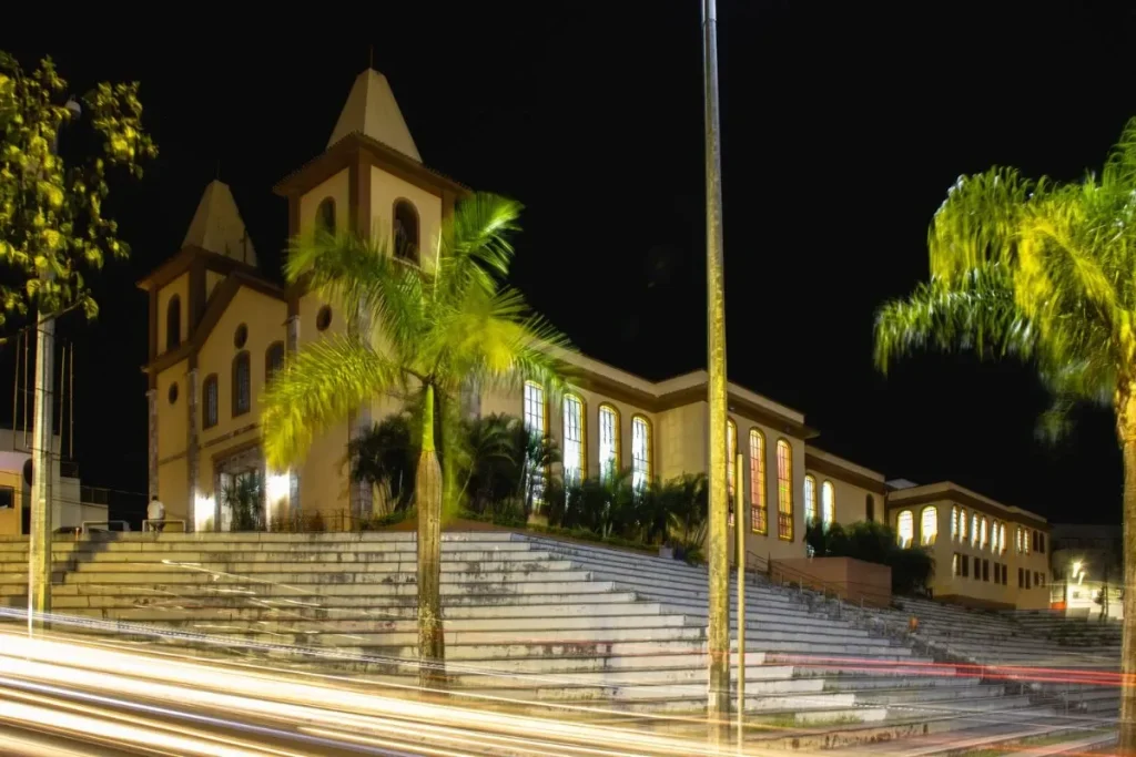 Fotografia noturna da Igreja Matriz de São Gonçalo em Contagem, MG. A igreja, de estilo colonial, está iluminada em tons quentes e localizada no topo de uma grande escadaria. Em primeiro plano, rastros de luz brancos e vermelhos de carros em movimento atravessam a imagem, uma técnica de longa exposição.