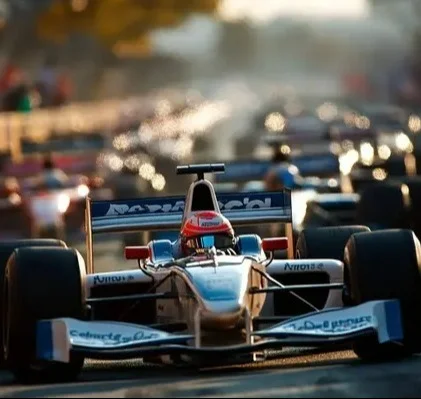 Visão frontal de um carro de Fórmula 1 com livery azul e branca, focado nitidamente em um grid de largada. O piloto, com capacete vermelho, está visível no cockpit. O fundo está muito desfocado, mostrando silhuetas de outros carros e um forte brilho de luzes quentes.