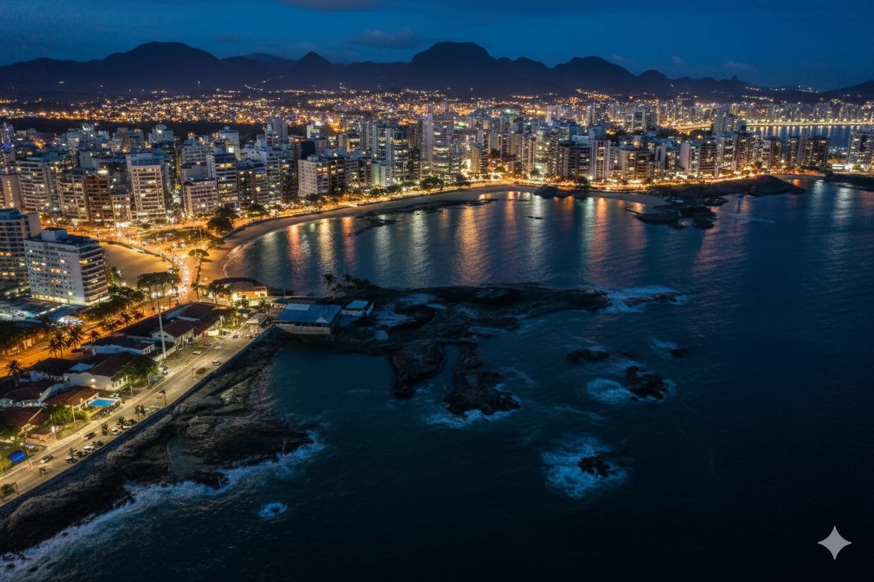 Vista aérea noturna da orla central de Guarapari, Espírito Santo. A imagem captura a vibrante iluminação da cidade que contorna a praia, com as montanhas ao fundo sob um céu crepuscular.