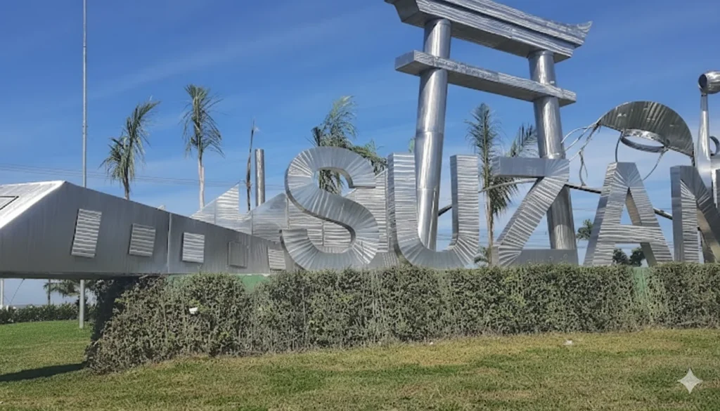 Uma imagem diurna de um grande monumento com a palavra "SUZANO" escrita em letras trateadas e tridimensionais, em um estilo moderno. As letras são altas e refletem a luz do sol, que brilha intensamente de um céu azul claro e sem nuvens. Elementos decorativos associados à cultura japonesa estão integrados na estrutura, incluindo um portão torii de metal prateado que se eleva sobre uma das letras e formas curvas que lembram as dobras de um origami ou um peixe koi estilizado em outras partes do monumento. Palmeiras esguias e algumas folhagens baixas pontuam o fundo, enquanto na base do monumento há um canteiro de arbustos baixos e densos, e na frente, um gramado verde e bem cuidado. A cena transmite uma sensação de brilho e orgulho cívico.