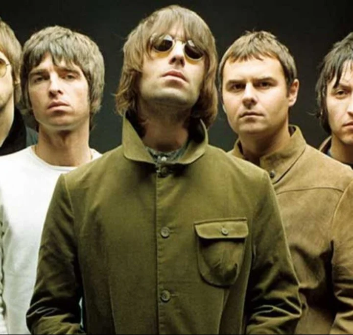 Foto colorida da banda Oasis em um retrato de grupo. O vocalista Liam Gallagher está no centro e em primeiro plano, usando óculos de sol redondos e uma jaqueta verde. Atrás dele estão os outros membros da banda, incluindo Noel Gallagher, todos com expressões sérias contra um fundo escuro.