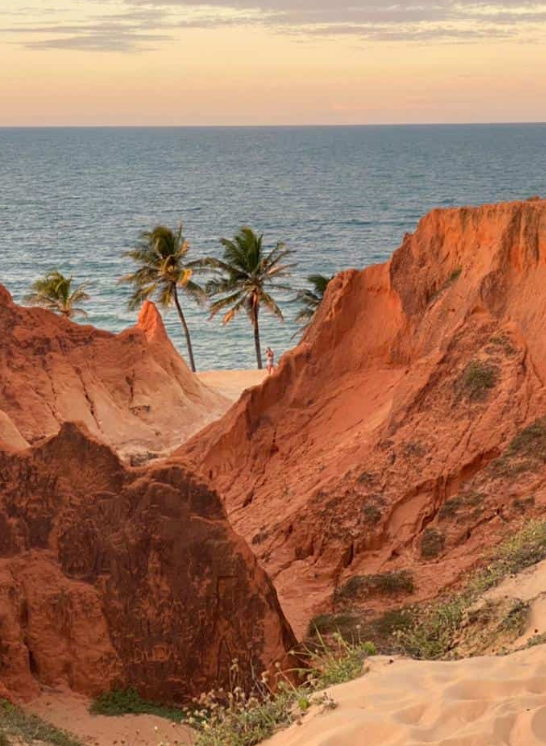 Fotografia de paisagem litorânea em Beberibe, Ceará, exibindo imponentes falésias avermelhadas em primeiro plano, com coqueiros verdes na faixa de areia e o mar azul ao fundo sob um céu nublado ao entardecer.
