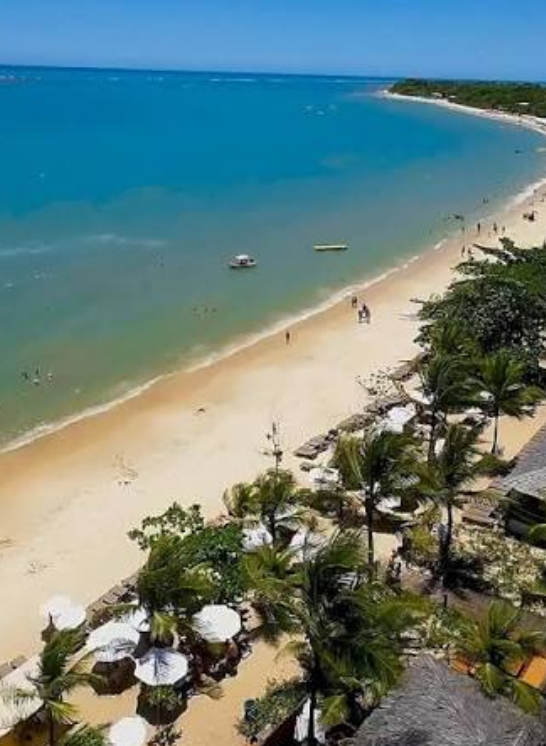 Vista aérea da praia em frente ao La Torre Resort All Inclusive em Porto Seguro, mostrando o mar em tons de azul e verde, faixa de areia clara, coqueiros e a estrutura de sombreiros brancos do resort à beira-mar.
