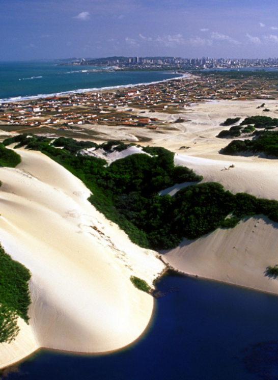 Vista aérea panorâmica de Natal, Rio Grande do Norte, destacando em primeiro plano uma lagoa azul cercada por dunas de areia branca e vegetação nativa, com a cidade e a costa litorânea estendendo-se ao fundo sob um céu ensolarado.