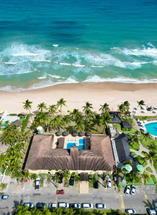 Fotografia aérea vertical do Atlântico Sul Hotel em Porto de Galinhas, mostrando a fachada do hotel, sua área de piscina interna, carros estacionados na rua frontal, e uma ampla faixa de areia clara que se conecta ao mar azul com ondas cristalinas.