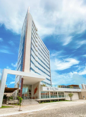 Fotografia contrapicada de um hotel moderno e alto em formato de cunha, pintado de branco com faixas de janelas escuras, apresentando na base o letreiro "Bali Mar Hotel", sob um céu azul intenso com nuvens.