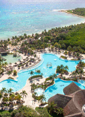 Vista aérea de um amplo resort tropical mostrando uma imensa piscina azul clara, construções com telhados de palha, uma praia de areia branca e o oceano turquesa ao fundo, cercados por vegetação densa e verde