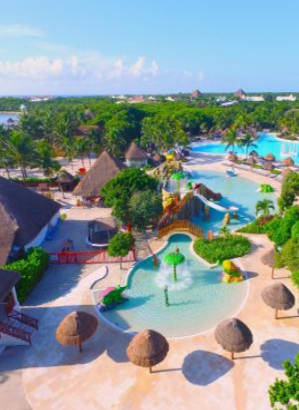 Vista aérea de uma área de piscina infantil em um resort tropical, mostrando brinquedos aquáticos coloridos, um pequeno escorregador e fontes em forma de cogumelo, com uma grande piscina azul, construções com telhado de palha e floresta densa ao fundo.