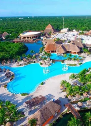 Vista aérea de um grande resort tropical com uma extensa piscina azul clara, cercada por espreguiçadeiras, construções com telhados de palha e palmeiras, tendo uma densa floresta verde se estendendo até o horizonte ao fundo.