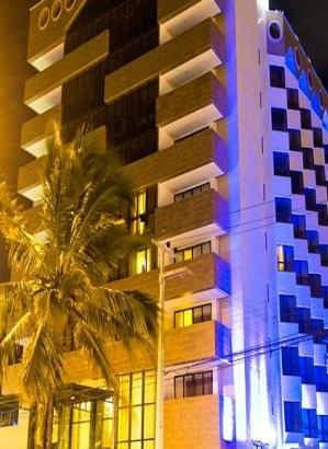 Fotografia noturna em ângulo contrapicado de um edifício alto, com sacadas salientes em tons de bege e uma fachada lateral intensamente iluminada em azul elétrico, com uma grande palmeira em destaque no primeiro plano à esquerda.
