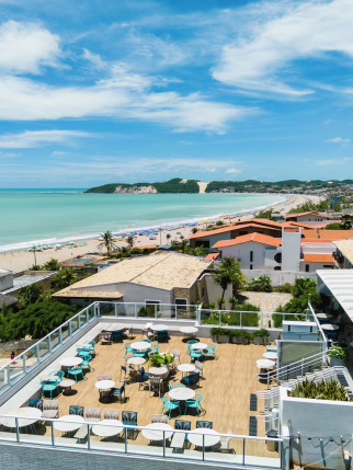 Vista elevada de um terraço ao ar livre com mesas brancas e cadeiras azuis em primeiro plano, com vista para uma praia, mar azul-esverdeado e uma encosta verde com uma grande duna de areia ao fundo, sob um céu azul.