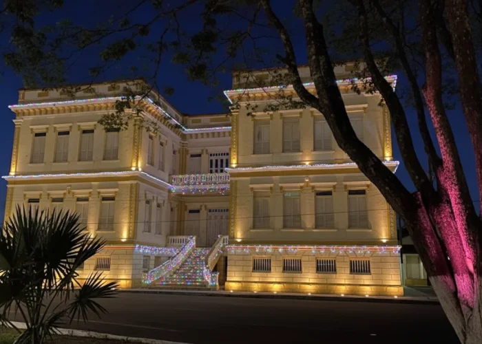 Uma foto noturna da Prefeitura de Bom despacho, um prédio de dois andares com arquitetura clássica e paredes em tom de creme. O edifício está decorado com luzes de Natal coloridas em cascata nas varandas e na escadaria principal, que leva à entrada do segundo andar. Holofotes na base iluminam a fachada. À direita, uma árvore com o tronco e os galhos são iluminados por uma luz rosa. O céu noturno está em um tom de azul escuro.