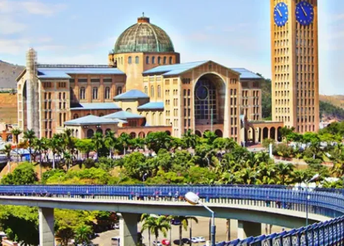 Vista panorâmica da Basílica do Santuário Nacional de Aparecida, um imenso templo de tijolos com grandes cúpulas e uma alta torre de sino, cercada por jardins e uma grande praça sob um céu azul.