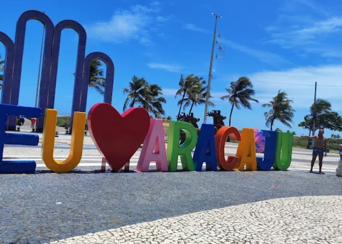 Letreiro turístico colorido "Eu Amo Aracaju" na Orla de Atalaia, em Aracaju. Ao fundo, veem-se os Arcos da Orla, coqueiros e um céu azul com algumas nuvens.