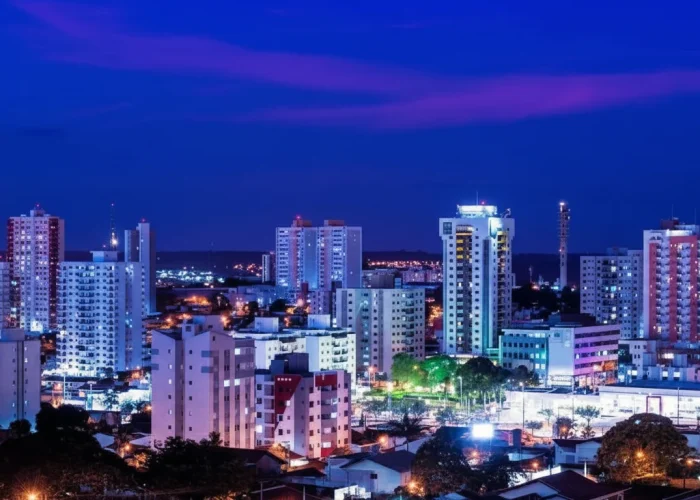 Uma imagem noturna deslumbrante de um horizonte urbano. Edifícios de várias alturas, alguns com múltiplos andares e janelas iluminadas, dominam o centro da imagem. As luzes dos edifícios e da rua emitem um brilho predominantemente branco e azulado, embora alguns pontos exibam tons rosados e roxos, especialmente visíveis nas luzes dos prédios mais altos e no céu. O céu acima da cidade é de um azul profundo, quase índigo, com nuvens alongadas e escuras tingidas de rosa e roxo, sugerindo o final do crepúsculo. Na parte inferior da imagem, há telhados de casas e a copa de algumas árvores, também iluminadas por pontos de luz amarela. A imagem transmite uma sensação de vastidão e movimento, com a cidade se estendendo para o horizonte sob um céu dramático.