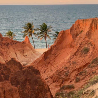 Fotografia de paisagem litorânea em Beberibe, Ceará, exibindo imponentes falésias avermelhadas em primeiro plano, com coqueiros verdes na faixa de areia e o mar azul ao fundo sob um céu nublado ao entardecer.