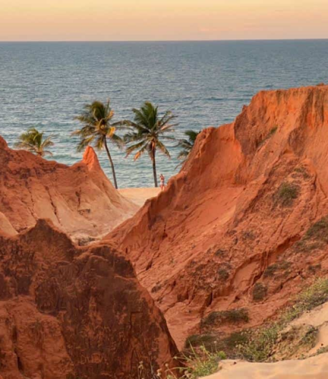 Fotografia de paisagem litorânea em Beberibe, Ceará, exibindo imponentes falésias avermelhadas em primeiro plano, com coqueiros verdes na faixa de areia e o mar azul ao fundo sob um céu nublado ao entardecer.