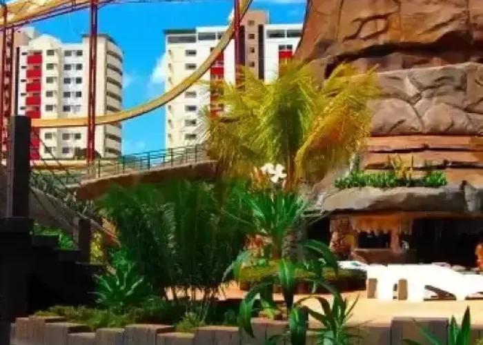 Rio lento no diRoma Acqua Park cercado por jardins tropicais, com toboágua amarelo e estrutura rochosa com cascata, e prédios ao fundo sob um céu azul com nuvens.