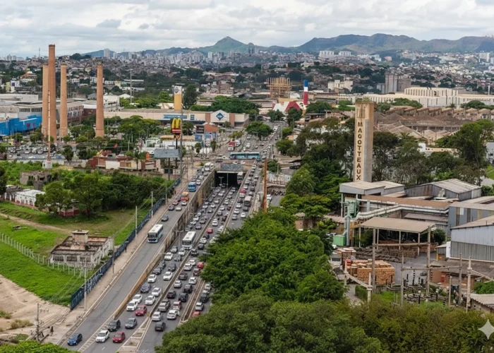 Vista aérea da Rodovia BR-381 na altura da Cidade Industrial, em Contagem, com trânsito intenso. A paisagem inclui marcos industriais, como três chaminés altas de tijolos. Ao fundo, a cidade de Belo Horizonte se estende até a Serra do Curral sob um céu nublado.