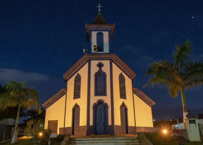 "Uma igreja colonial branca com detalhes em azul-escuro, incluindo uma torre central com uma cruz, é vista à noite. A fachada é iluminada por luzes amarelas quentes, destacando sua arquitetura. Duas palmeiras ladeiam a igreja sob um céu noturno azul-marinho pontilhado de estrelas, com escadarias e vegetação em primeiro plano."