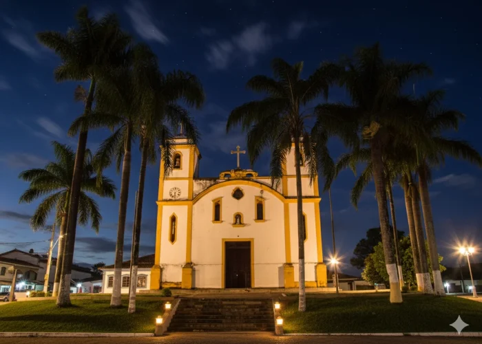 "Uma igreja colonial branca com detalhes em amarelo, duas torres sineiras, um relógio em uma delas e uma cruz no topo da fachada, é vista à noite. Palmeiras altas emolduram a estrutura, e a iluminação noturna realça a arquitetura e os degraus que levam à entrada. O céu noturno é azul-escuro com algumas nuvens e estrelas visíveis, e há postes de luz ao redor."