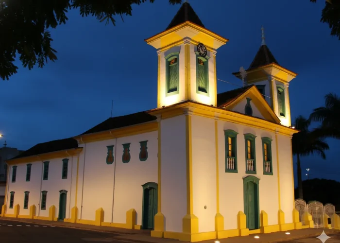 Fotografia noturna da Igreja Matriz de São Gonçalo, localizada em São Gonçalo do Pará, MG. A igreja de arquitetura colonial, com uma torre central e fachada em tons de amarelo, está iluminada por luzes douradas que a destacam contra o céu azul-escuro. A foto é tirada a partir da praça central em frente ao monumento.