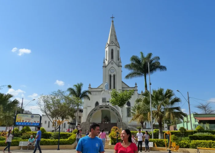 Uma imagem vibrante de uma igreja branca de arquitetura gótica, com uma torre alta e pontiaguda no centro, sob um céu azul claro com algumas nuvens esparsas. Em frente à igreja, há uma praça gramada com arbustos bem cuidados, palmeiras e algumas pessoas caminhando. Bancos brancos e equipamentos de ginástica amarelos são visíveis na praça. Um casal caminha em primeiro plano no centro. À esquerda, um banner colorido está pendurado perto de outras árvores e construções.