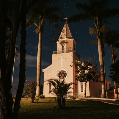 Emoldurada por palmeiras imponentes e sob um céu noturno estrelado, a igreja de tom pêssego irradia uma serenidade acolhedora. A iluminação suave realça seus detalhes arquitetônicos e a beleza tranquila de sua rosácea, convidando à contemplação noturna.