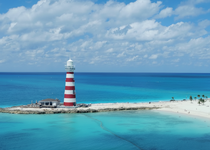 Vista panorâmica de um farol com listras vermelhas e brancas na ponta de uma ilha. O farol está cercado por um mar de águas cristalinas de cor turquesa, com uma praia de areia branca e céu azul com nuvens ao fundo.