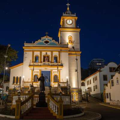 A histórica Igreja Matriz de São Lourenço da Serra, em São Paulo, destaca-se em uma bela noite de céu limpo. A iluminação artificial realça os detalhes de sua arquitetura colonial, desde a torre do relógio até a estátua de seu padroeiro, criando uma atmosfera serena e acolhedora no centro da cidade.