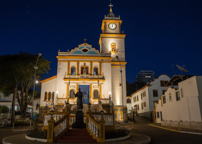 Fotografia noturna da Igreja Matriz de São Lourenço da Serra. A igreja de estilo colonial, pintada de branco com detalhes em amarelo e azul, está totalmente iluminada contra um céu escuro e estrelado. Em frente, há uma praça circular com uma estátua em um pedestal. A torre do relógio da igreja também está acesa.