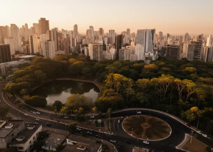Vista aérea do Lago das Rosas e do Parque Zoológico de Goiânia ao entardecer, com prédios da cidade ao fundo e uma rotatória com jardim ornamental em primeiro plano, sob um céu alaranjado.