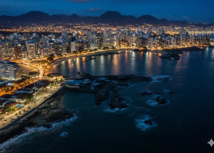 Foto aérea e noturna da cidade de Guarapari. Em primeiro plano, um trecho de costa rochosa e uma avenida. Ao centro, a baía principal com sua faixa de areia em formato de arco, cercada por prédios altos e densamente iluminados. As luzes da cidade se refletem na água escura do mar. Ao fundo, a silhueta de uma cadeia de montanhas se desenha contra o céu azul-escuro do anoitecer.