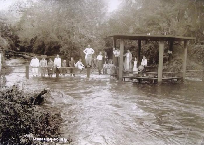 Fotografia antiga em preto e branco (sépia) de um grupo de pessoas, homens e mulheres, posando em uma ponte de madeira rústica e em um pequeno pavilhão coberto sobre um rio corrente. A vegetação é densa ao redor. Na parte inferior, lê-se a inscrição "Lembrança de Irahy".