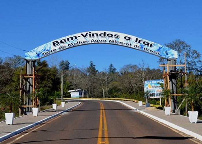Arco de boas-vindas na entrada de uma cidade com a inscrição "Bem-Vindos a Iraí Terra da Melhor Água Mineral do Brasil". O arco tem detalhes em azul que remetem à água. Abaixo, uma estrada asfaltada de duas mãos segue em direção à cidade. Há gramado, árvores e um céu azul claro. Um outdoor à direita também dá as boas-vindas.