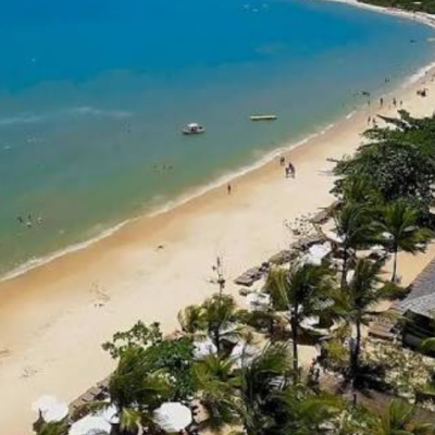 Vista aérea da praia em frente ao La Torre Resort All Inclusive em Porto Seguro, mostrando o mar em tons de azul e verde, faixa de areia clara, coqueiros e a estrutura de sombreiros brancos do resort à beira-mar.