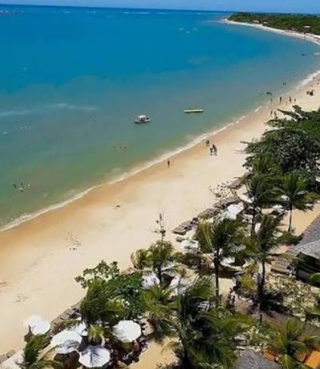 Vista aérea da praia em frente ao La Torre Resort All Inclusive em Porto Seguro, mostrando o mar em tons de azul e verde, faixa de areia clara, coqueiros e a estrutura de sombreiros brancos do resort à beira-mar.