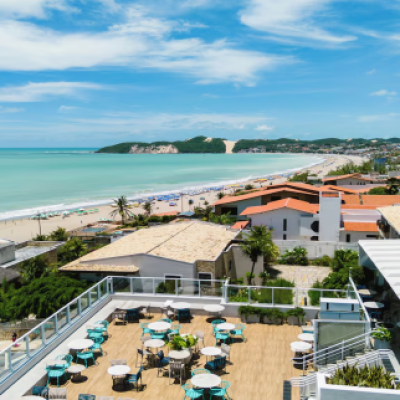 Vista elevada de um terraço ao ar livre com mesas brancas e cadeiras azuis em primeiro plano, com vista para uma praia, mar azul-esverdeado e uma encosta verde com uma grande duna de areia ao fundo, sob um céu azul.